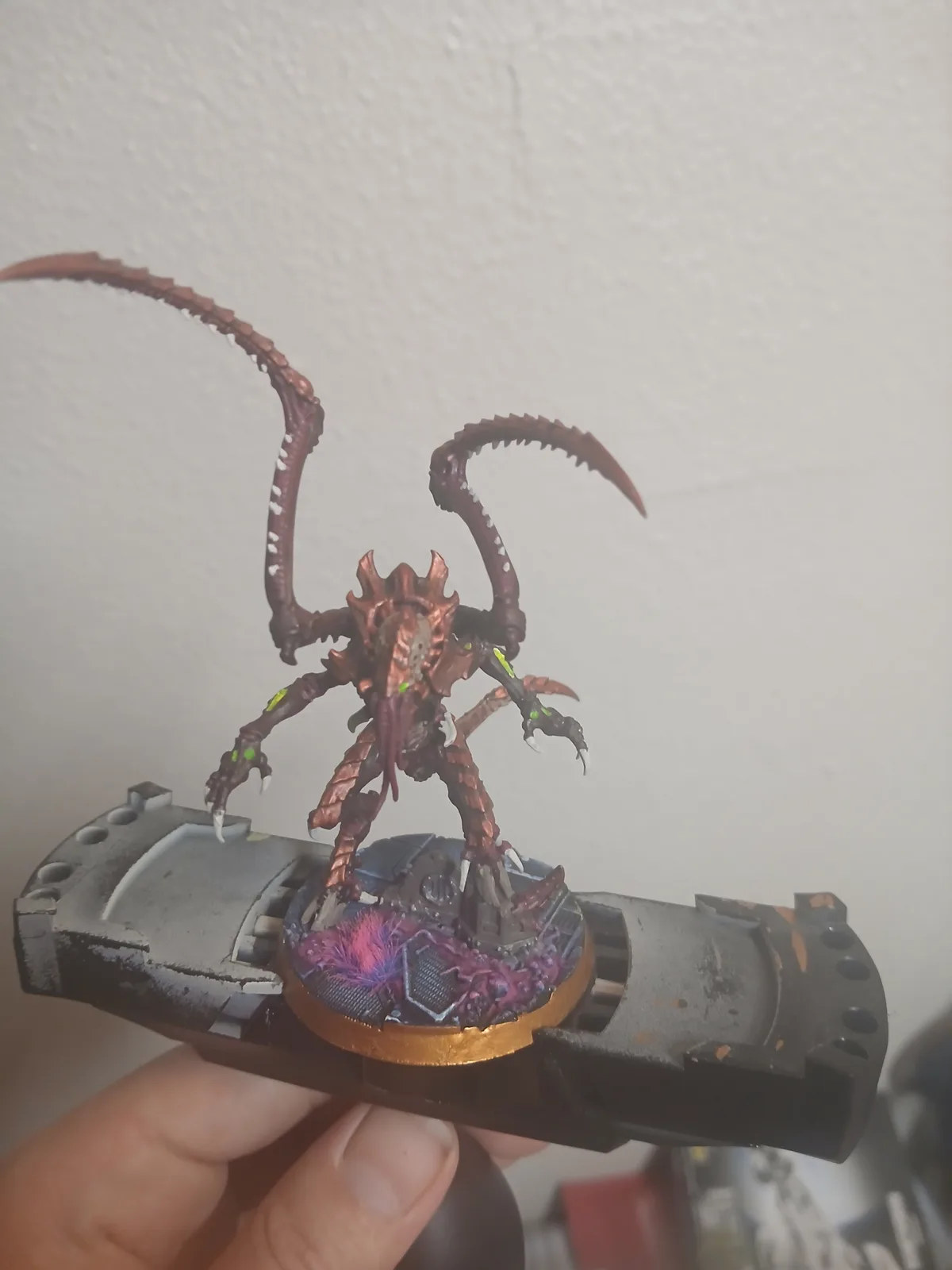 A hand holds a painted tabletop miniature of a grotesque, multi-limbed alien creature with long, curved, spiky appendages…