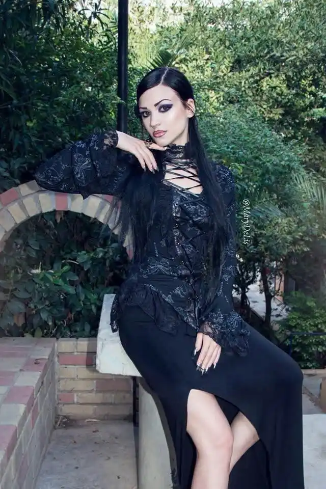 A woman with long black hair and dramatic gothic makeup poses outdoors in a black lace top with a lace-up neckline and a…