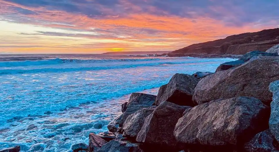 A vibrant sunset over a rocky coastline with crashing blue waves, overlaid with the text 'i'm not lazy, i'm in energy-saving…