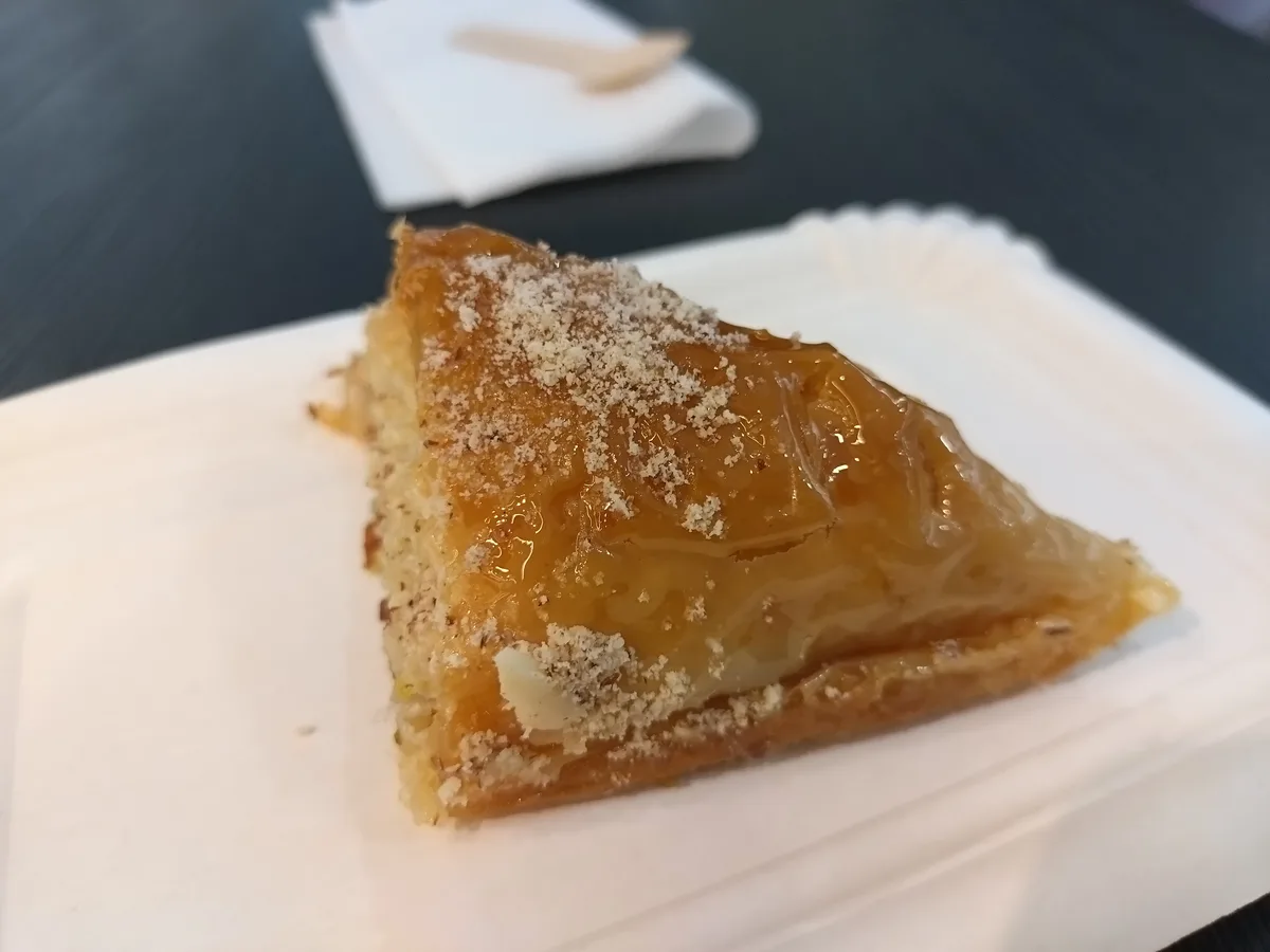 A close-up photo shows a triangular slice of baklava on a white paper plate, topped with a glossy golden syrup and dusted…