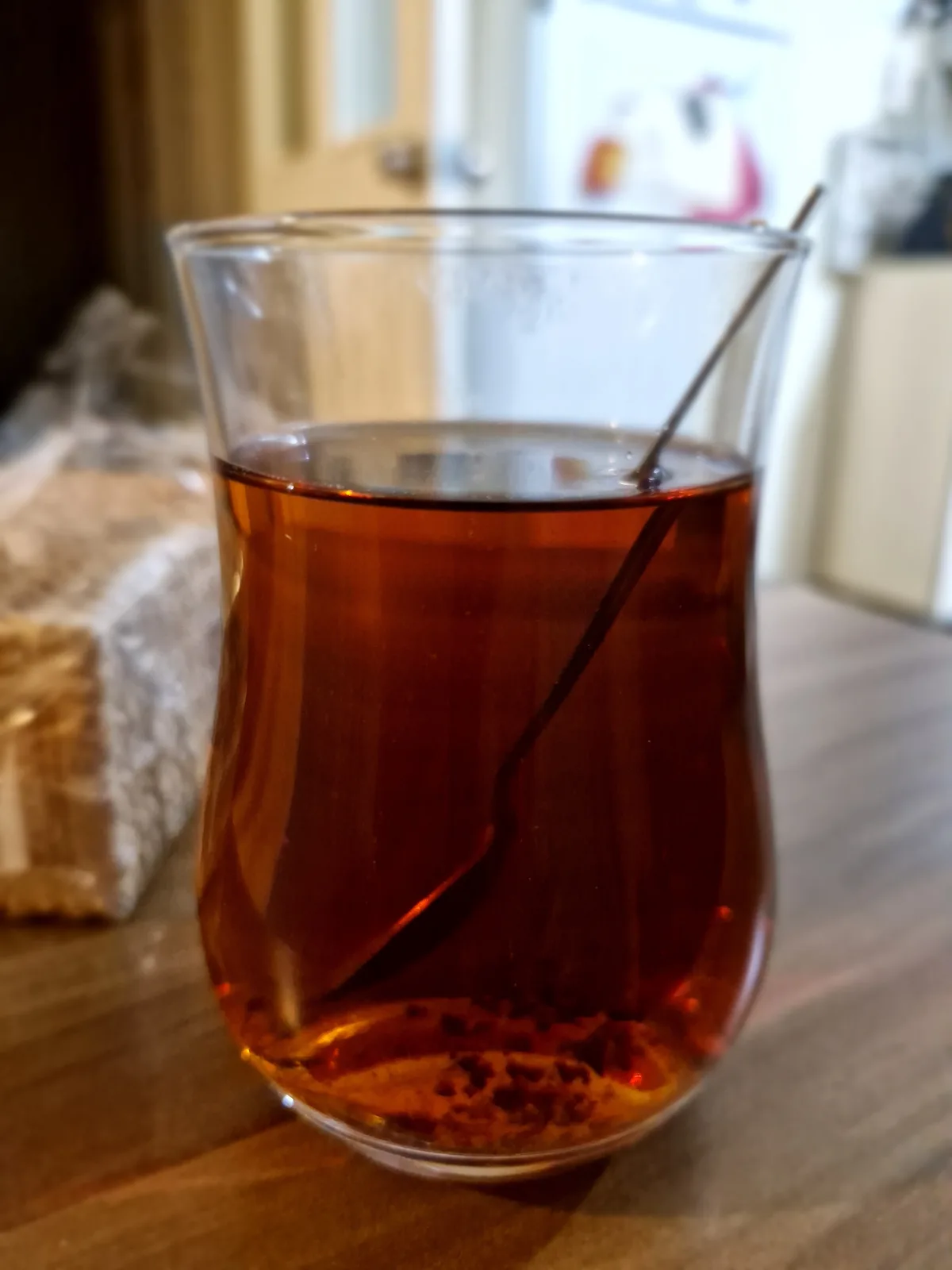 A close-up photo shows a clear glass teacup filled with dark amber tea, with a metal spoon resting inside. The cup sits on a…