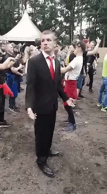 A man in a black suit and red tie stands stiffly in a muddy outdoor dance area, surrounded by people dancing. As the…