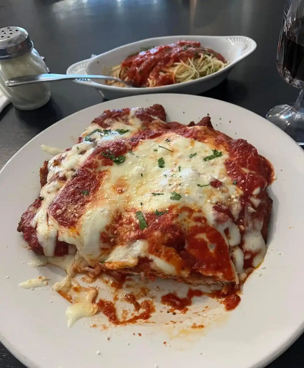 A close-up photo shows a large, messy plate of what appears to be chicken parm with melted cheese and tomato sauce…