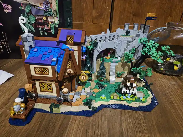 A completed LEGO set depicting a fantasy inn with a blue and brown roof, surrounded by greenery, a stone bridge, and a…