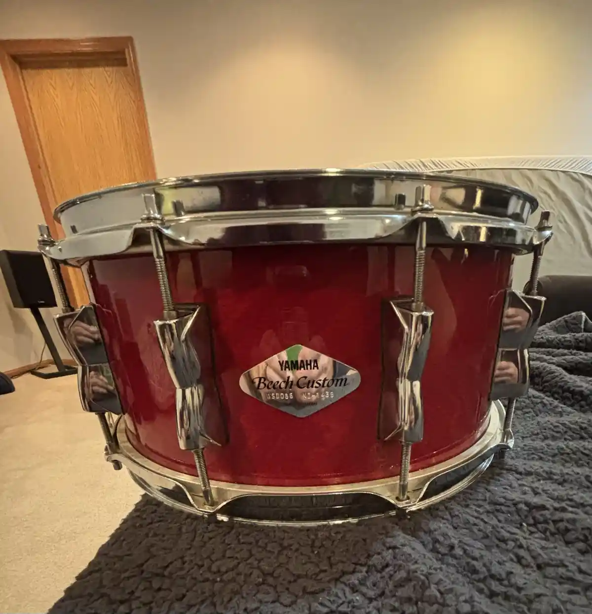 A glossy red Yamaha Beech Custom snare drum with chrome hardware sits on a dark gray textured blanket in a room, with a…
