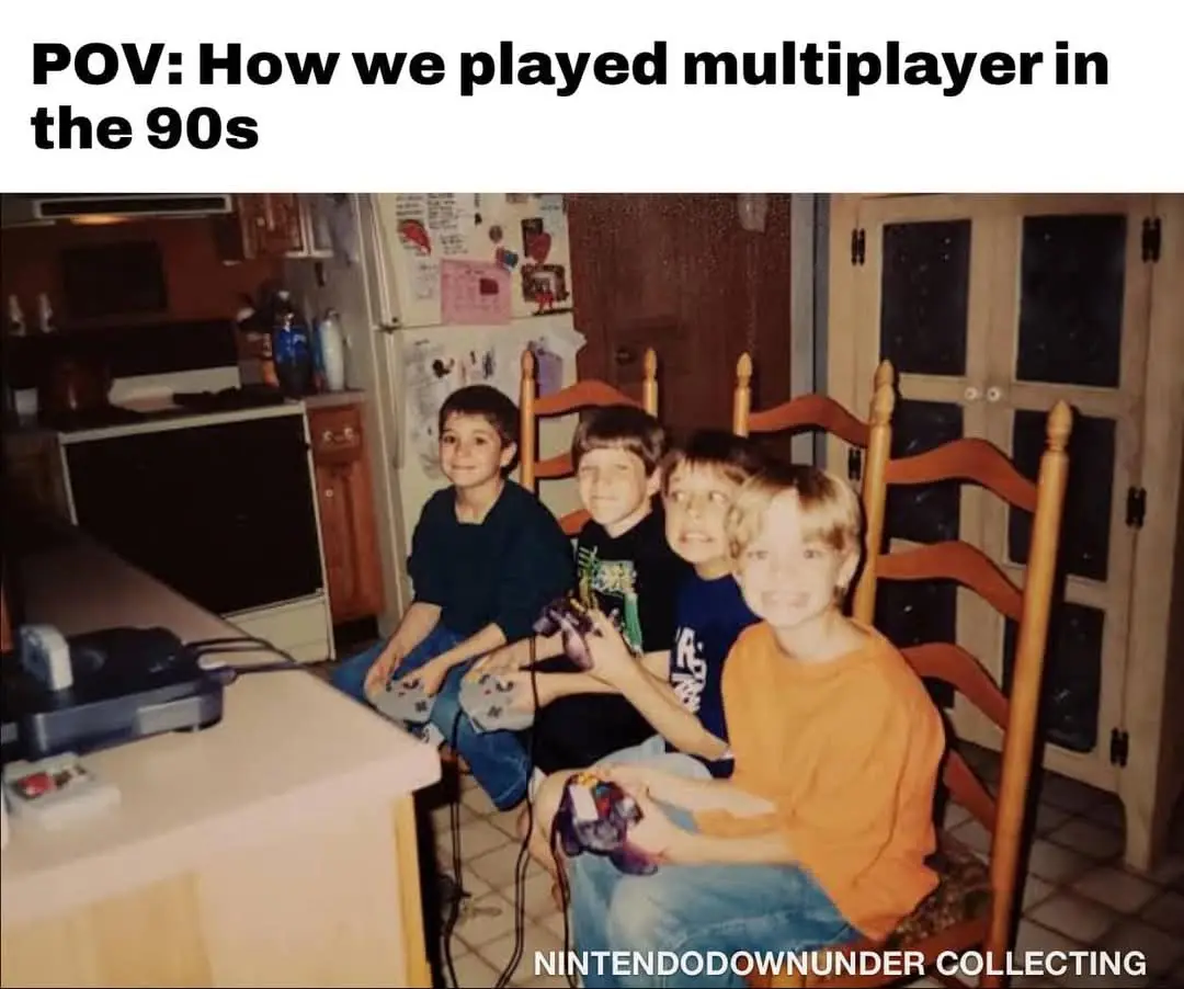 Four young boys sit on wooden chairs in a kitchen, each holding a purple Nintendo 64 controller, looking at the camera with…