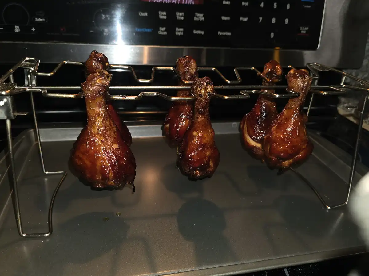 Six glazed chicken legs hang from a metal rack inside an oven, glistening with sauce, positioned above a baking tray. The…