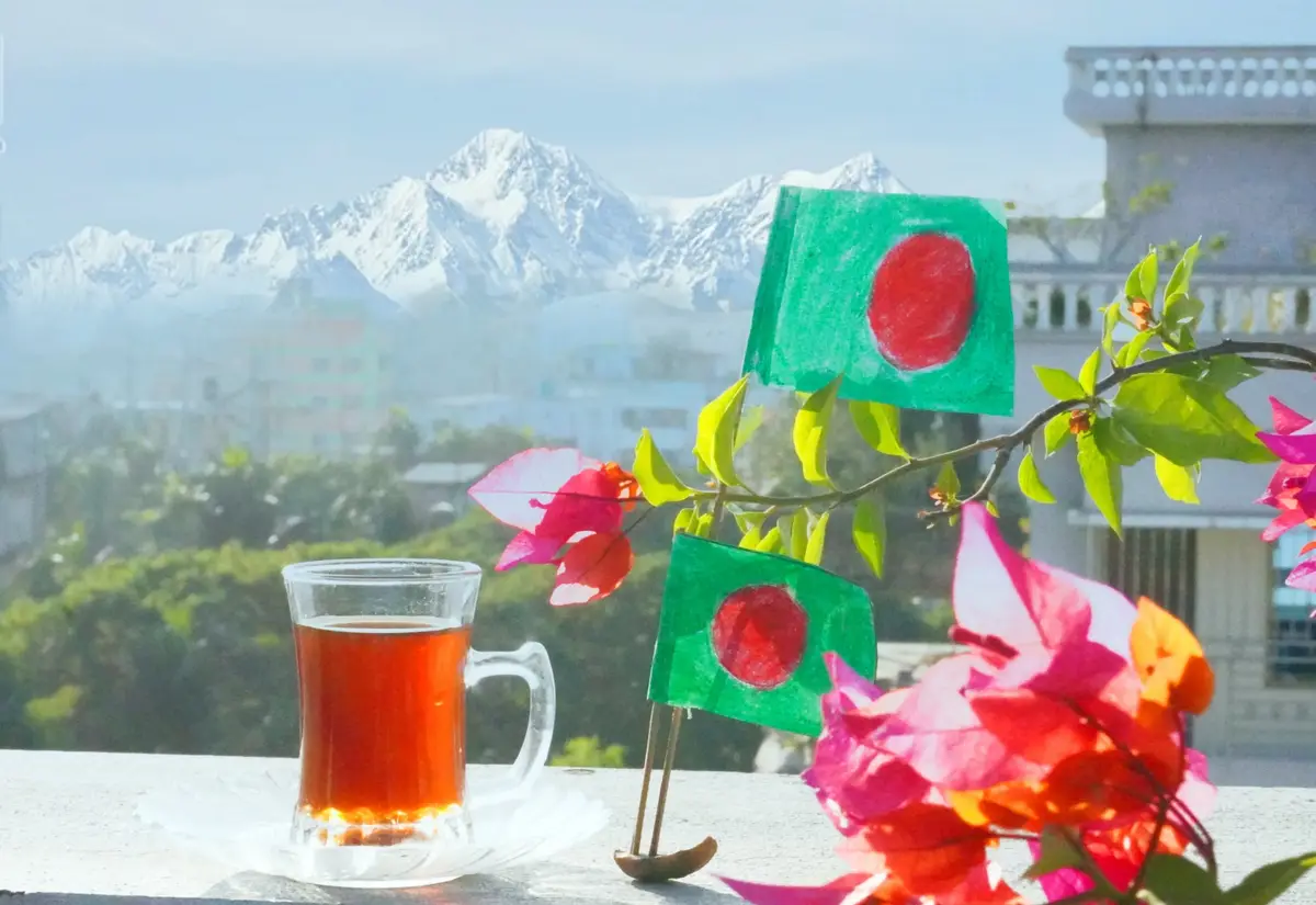 A glass mug of tea sits on a ledge with two small handmade Bangladesh flags on a stand beside it, framed by pink…
