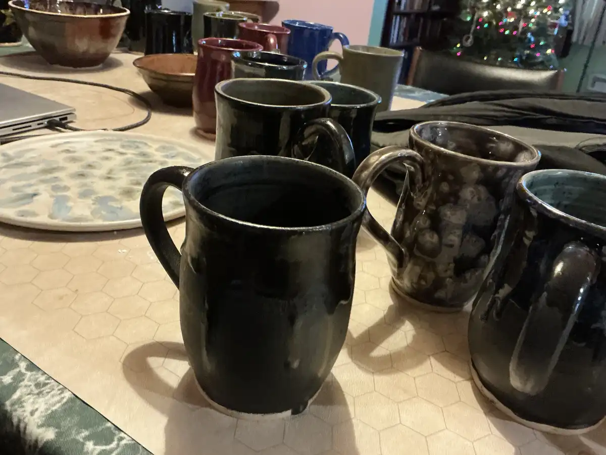A close-up photo shows a collection of handmade ceramic mugs, mostly dark and glossy, arranged on a hexagonal-patterned…