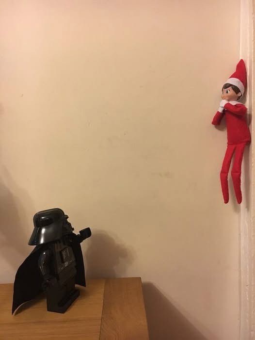 A red-clothed elf on the shelf is clinging to a wall corner, looking down at a LEGO Darth Vader figure on a wooden surface…