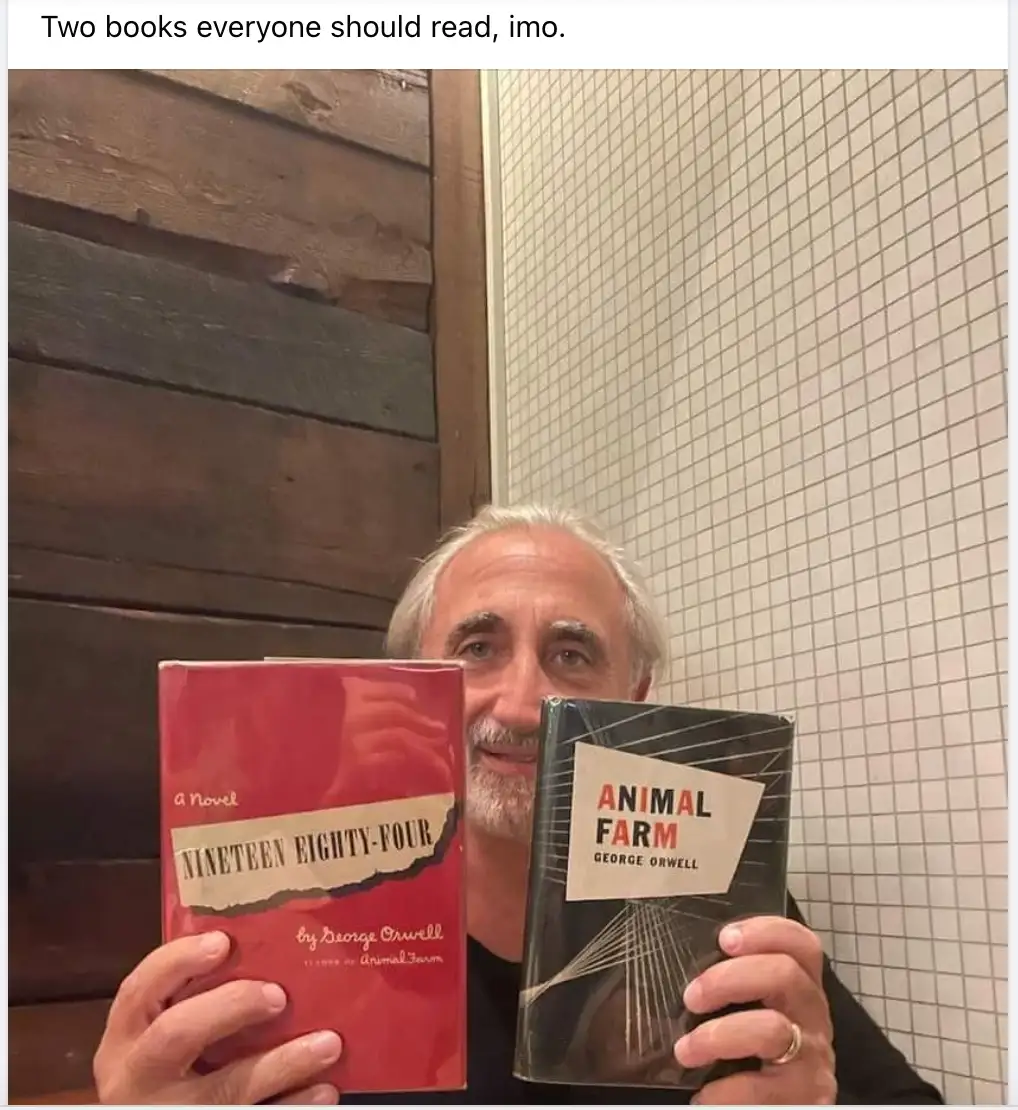 A man with gray hair and a beard holds up two books by George Orwell: 'Nineteen Eighty-Four' (red cover) and 'Animal Farm'…