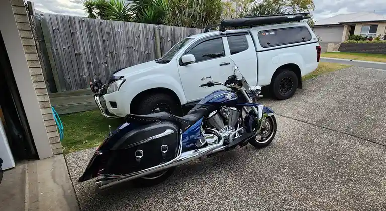 A shiny blue Victory motorcycle is parked in front of a white pickup truck with a canopy and roof rack, both vehicles are…