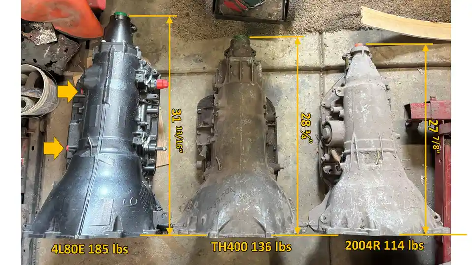 Three automotive transmissions are laid side-by-side on a concrete floor, each labeled with its model name, weight in…