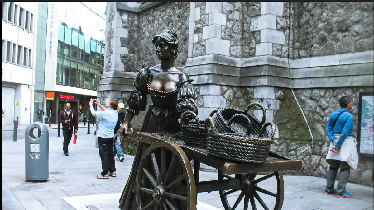 A bronze statue of a woman in period dress pushing a wooden cart loaded with woven baskets stands on a city sidewalk. In the…