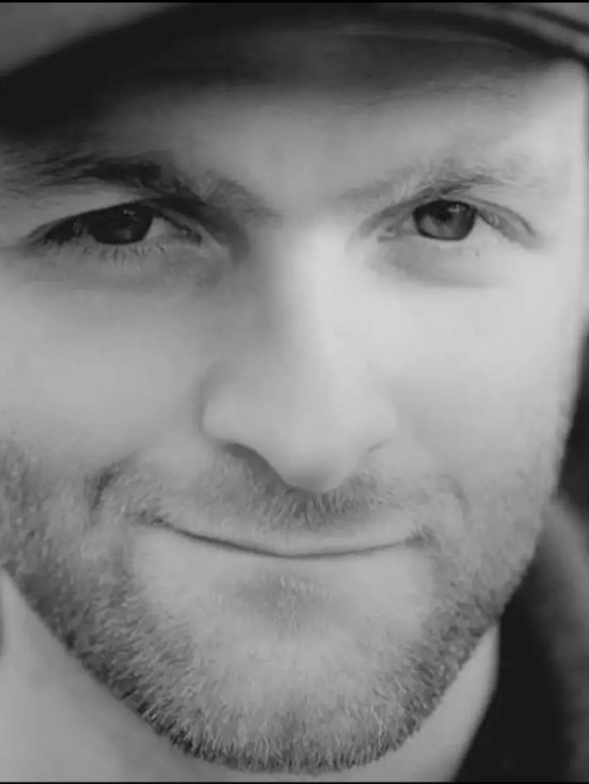 A black-and-white close-up of a man's face, showing his eyes, nose, and a slight smile. He wears a cap and has stubble. The…