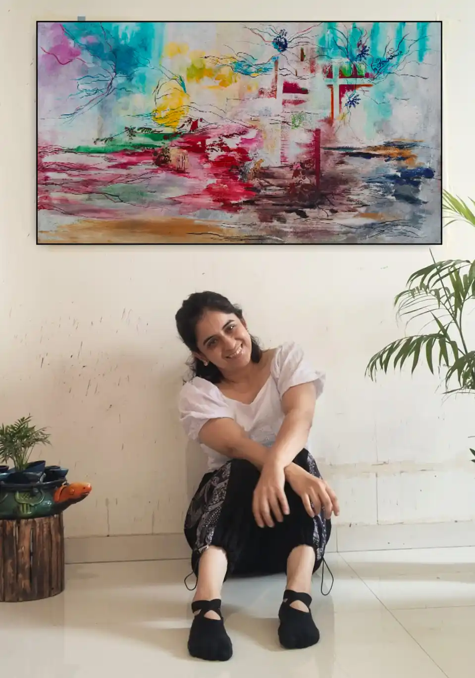 A woman sits on the floor smiling at the camera, positioned directly beneath a large, colorful abstract painting hanging on…