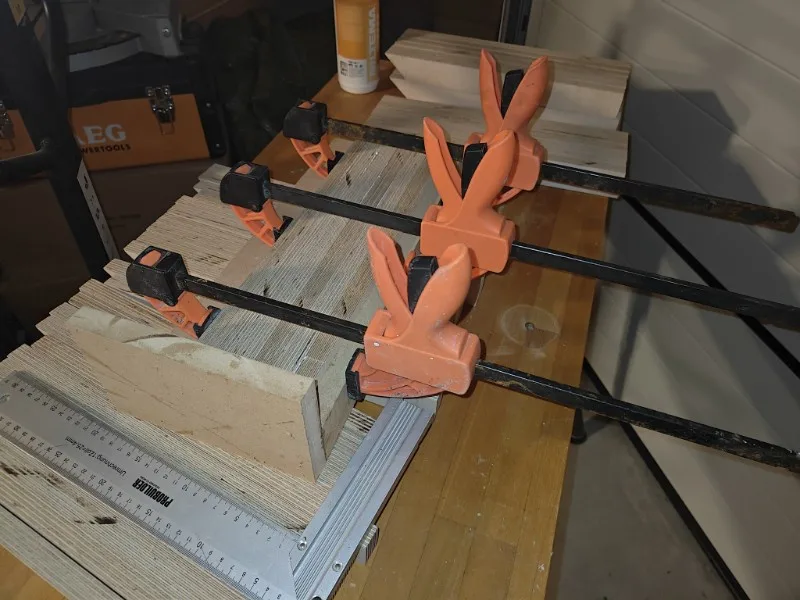 A woodworking scene shows multiple orange spring clamps holding stacks of wood together on a workbench. A metal ruler lies…