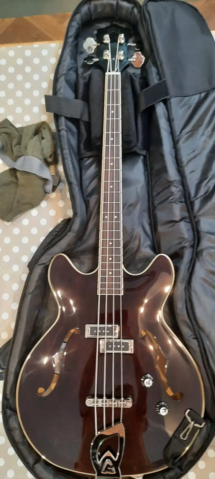 A dark brown, glossy electric bass guitar with four strings and f-holes is shown lying inside an open black padded case. The…