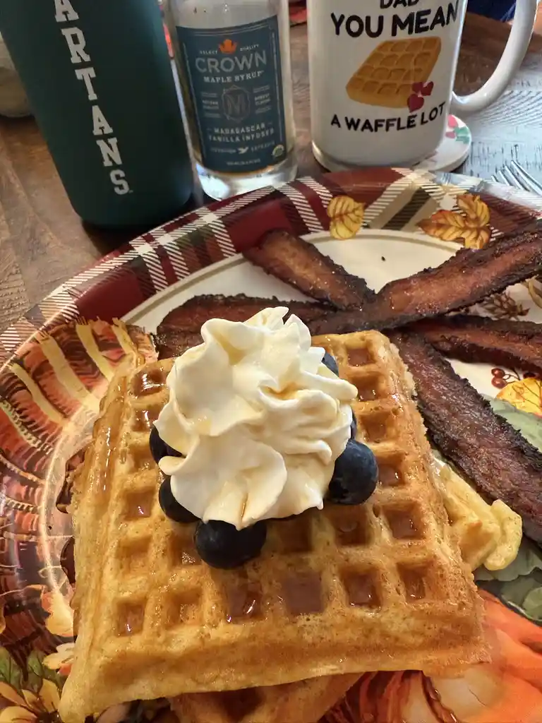 A close-up photo of a breakfast plate with a waffle topped with whipped cream and blueberries, surrounded by bacon strips…