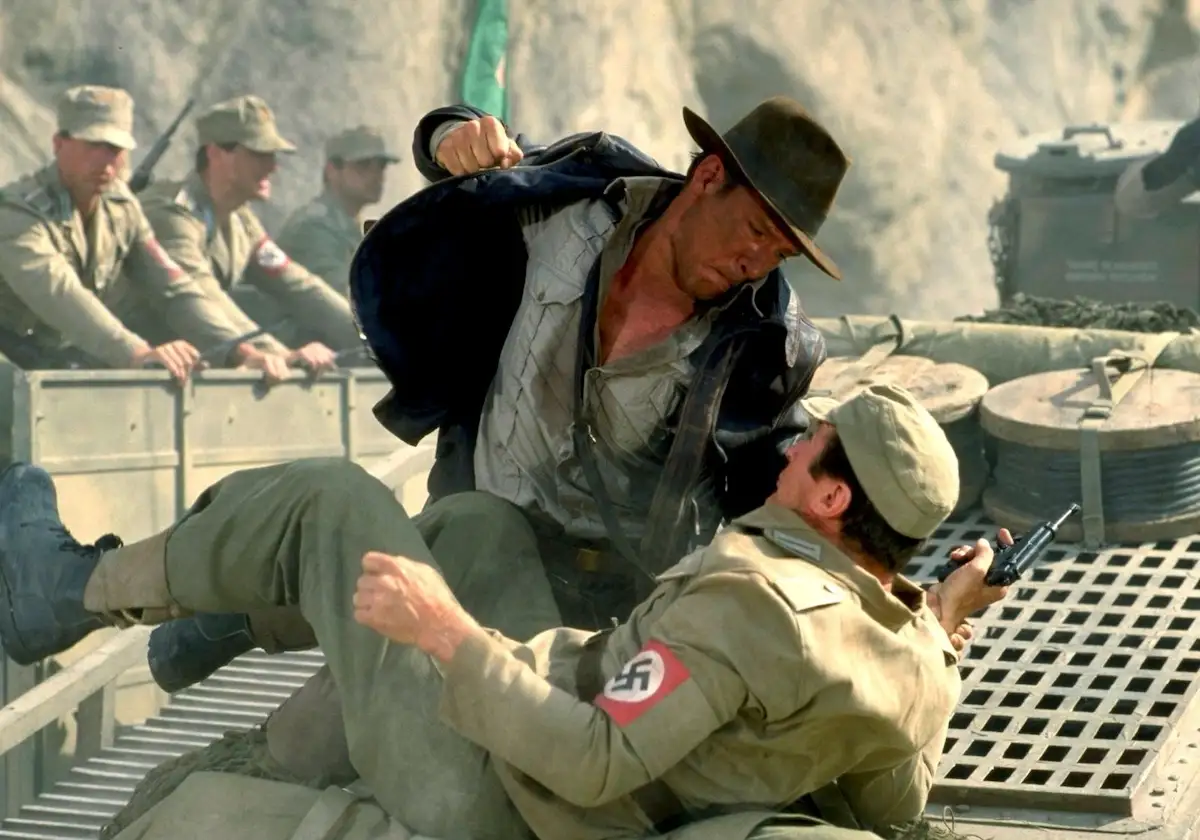 a man in a fedora and jacket is physically overpowering a soldier in a tan uniform with a swastika armband, who is holding a…