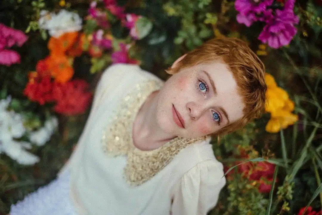 A young person with short red hair and blue eyes lies among colorful flowers, wearing a white top with a beaded collar. The…