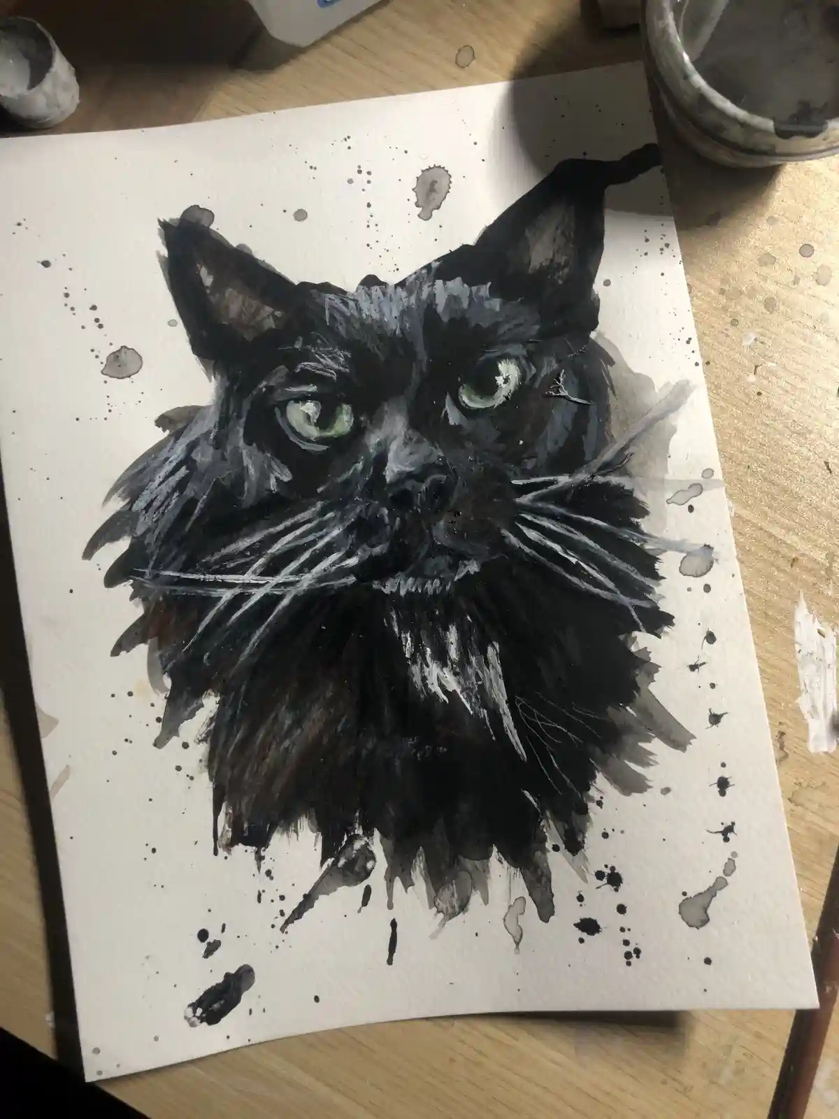 A watercolor painting of a black cat's head with green eyes and white whiskers on white paper, surrounded by paint…
