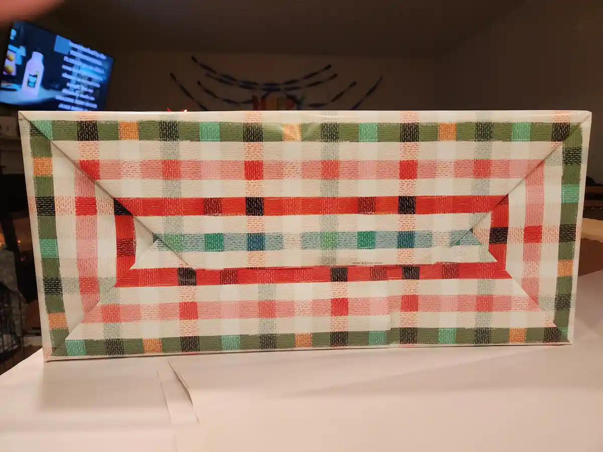 A rectangular gift box wrapped in colorful plaid-patterned paper sits on a white surface, with a 'happy birthday' banner…