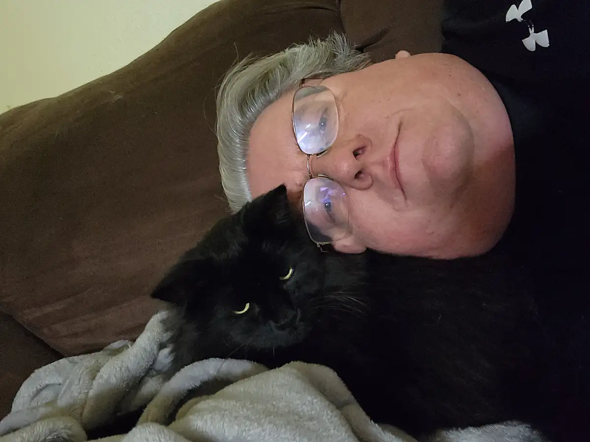A person with gray hair and glasses lies on a brown couch, smiling while a black cat rests its head on their chest. The cat…