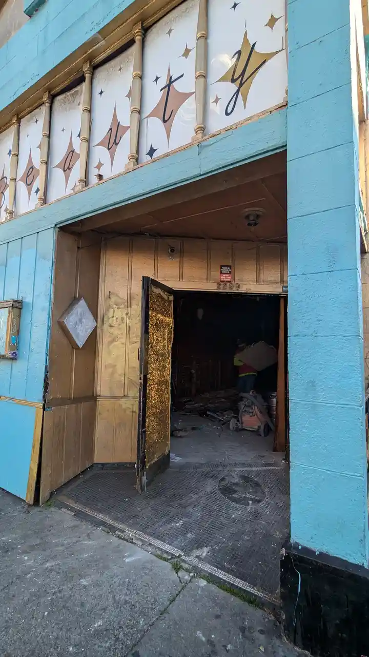 An exterior shot of a building with faded blue walls and a sign above the entrance that reads 'Beauty' in cursive script…