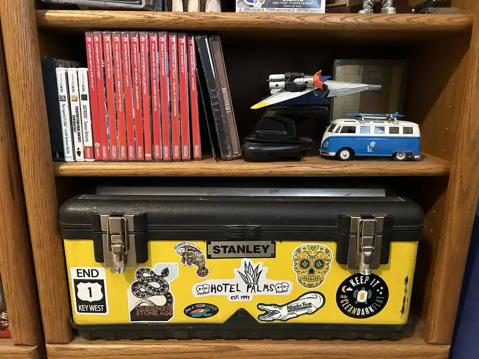 A yellow Stanley toolbox covered in stickers sits on a wooden shelf below a row of Nintendo Switch games and toy vehicles…