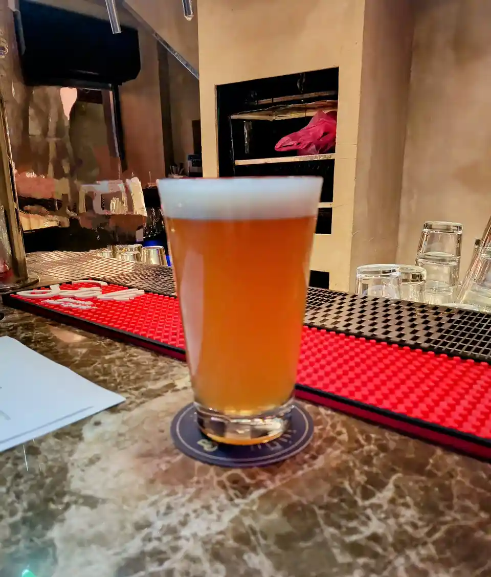 A tall glass of amber beer with a white head sits on a coaster on a marble bar, in front of a red bar mat and stacked…