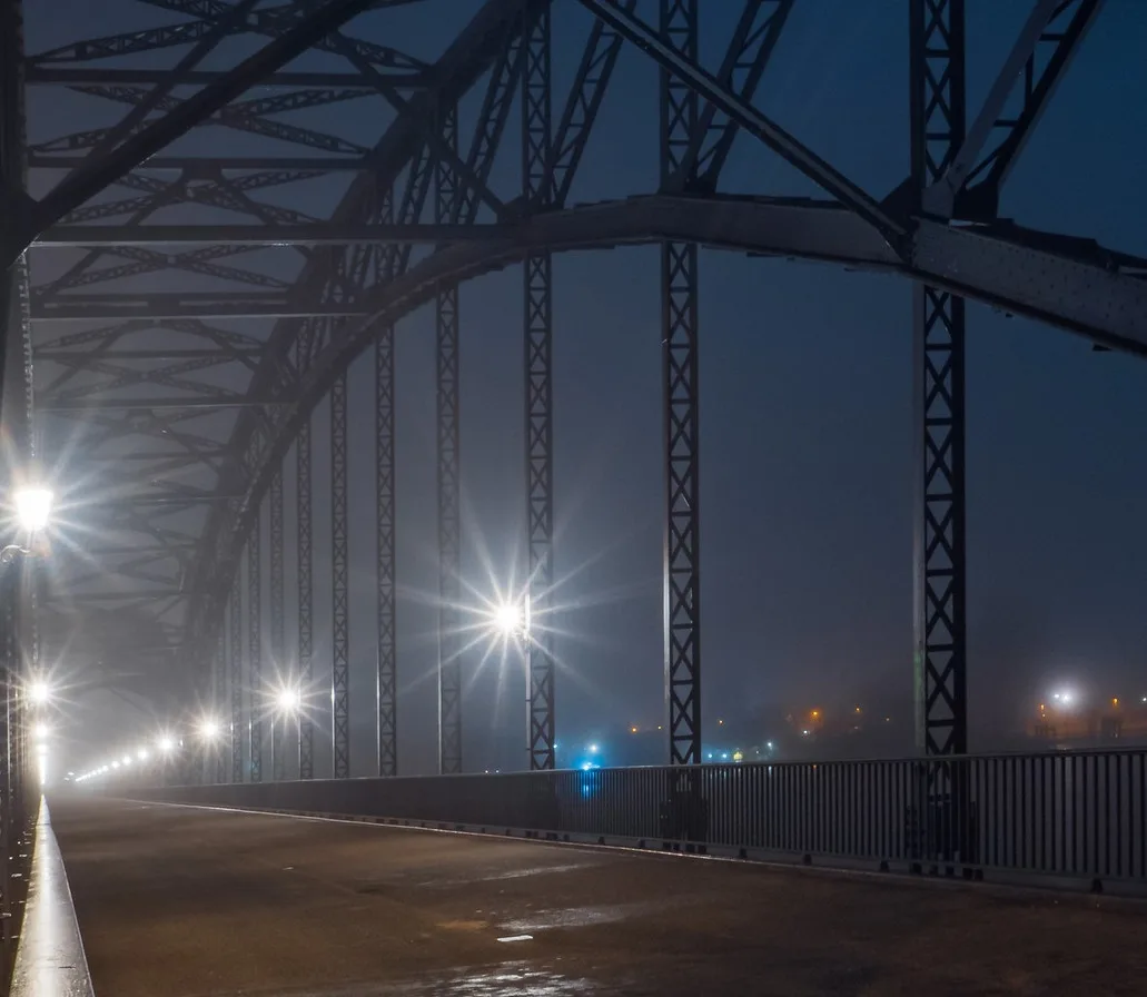 A nighttime photo of a steel truss bridge with bright streetlights casting starburst glares through fog, showing an empty…
