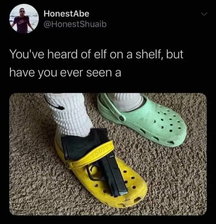 A close-up photo shows a person's feet on a carpet, one foot in a yellow Croc with a black handgun strapped to the top, the…