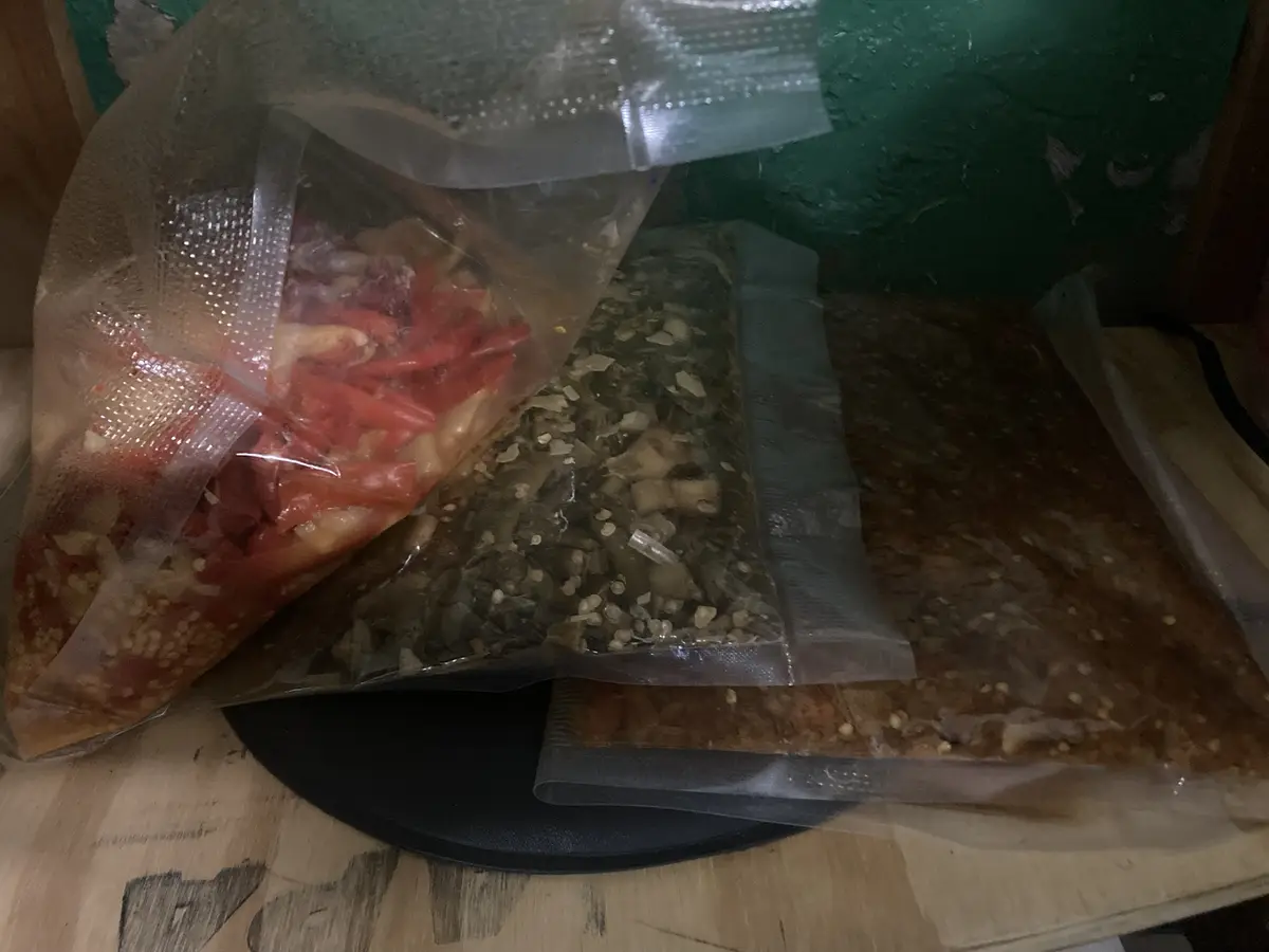 Three vacuum-sealed plastic bags filled with homemade pickled vegetables sit on a wooden surface. One bag contains red…