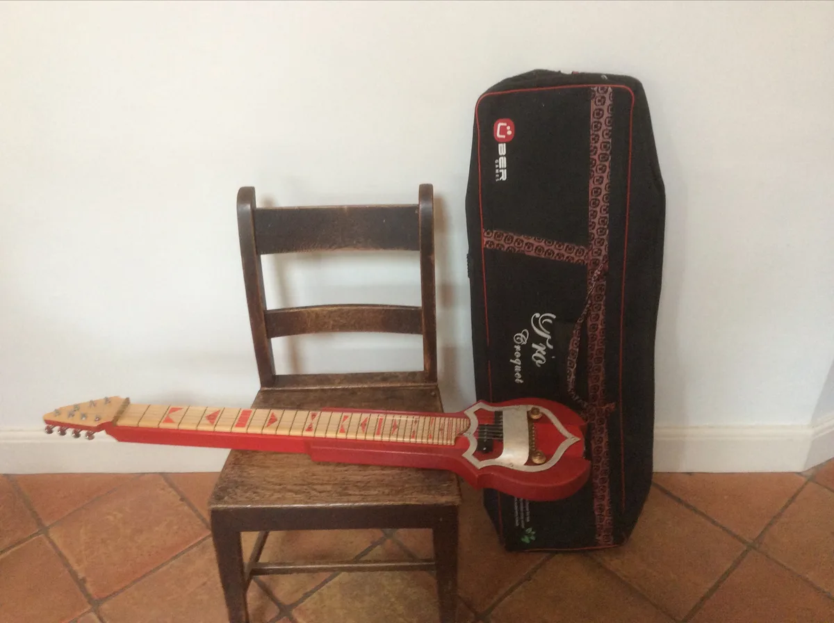 A red plastic guitar with red arrow-shaped fret markers rests horizontally on a wooden chair, next to a black guitar case…
