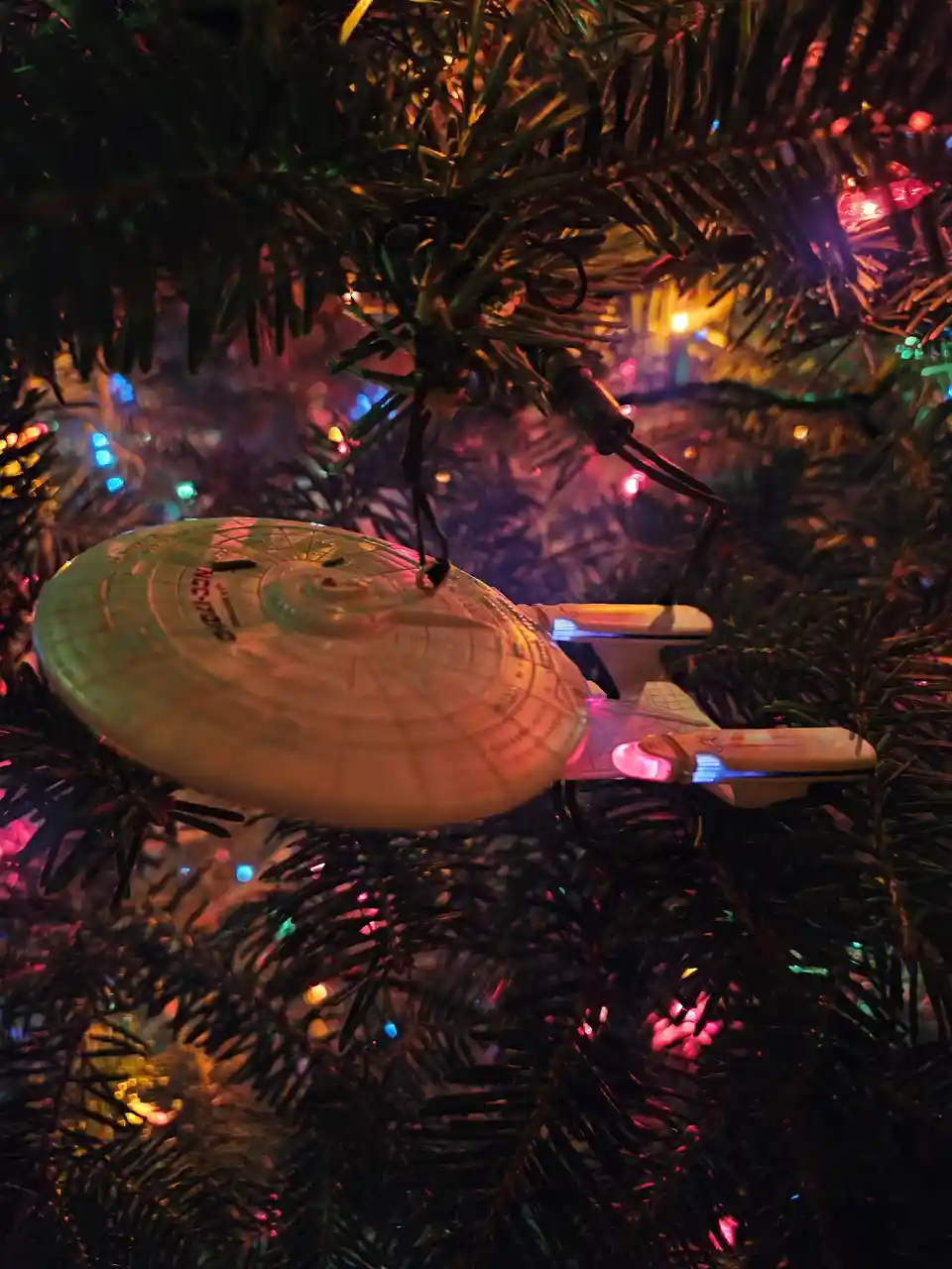 A Star Trek Enterprise model ornament hangs from a Christmas tree decorated with colorful lights, creating a festive sci-fi…