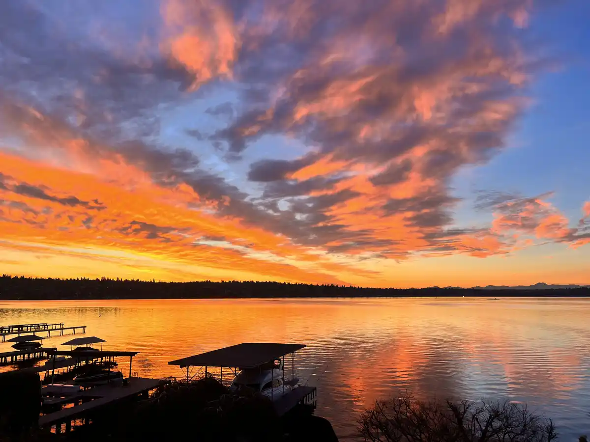 A serene sunset over a calm lake with docks and boats silhouetted in the foreground, featuring vibrant orange, pink, and…