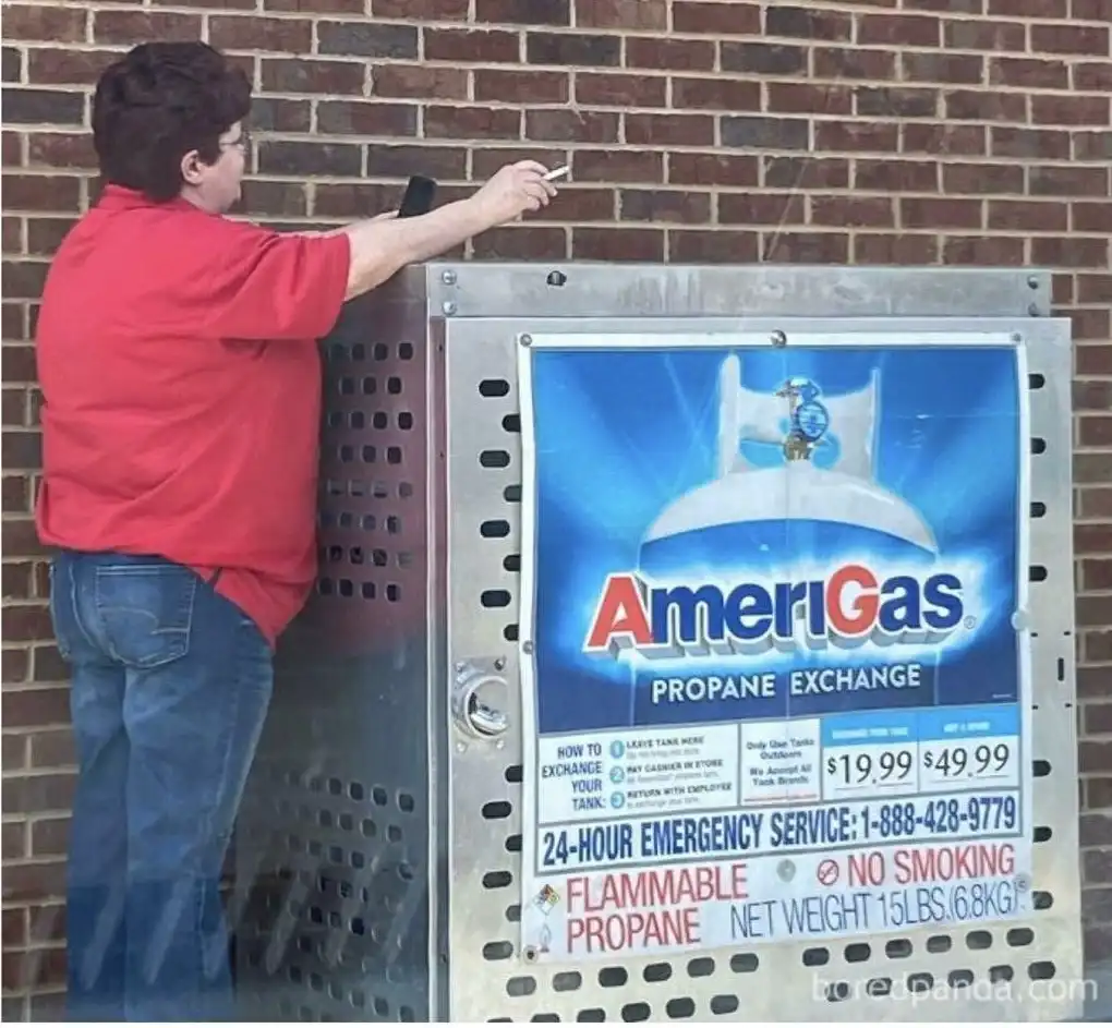 A person in a red shirt and jeans stands beside a metal propane exchange station with an 'AmeriGas' sign, holding a…