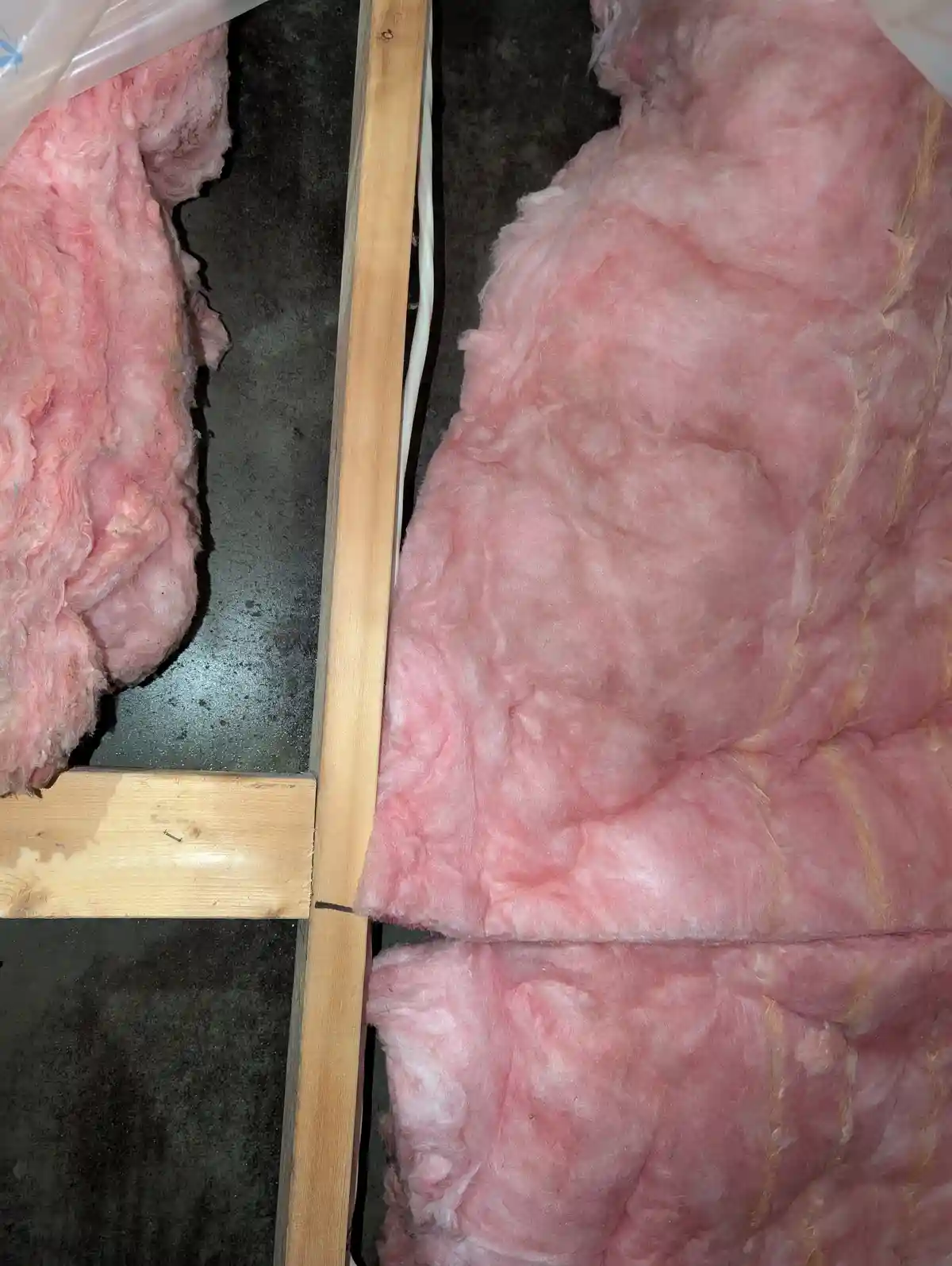 A close-up photo shows pink fiberglass insulation filling the space between wooden studs in a wall cavity, with a small gap…