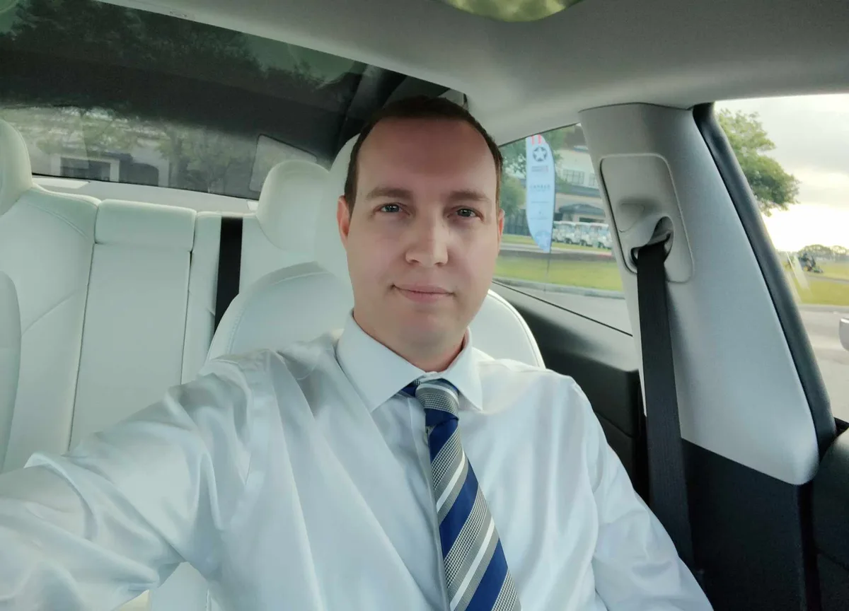 A man in a white shirt and blue striped tie sits in the driver's seat of a car with white leather interior, taking a selfie…