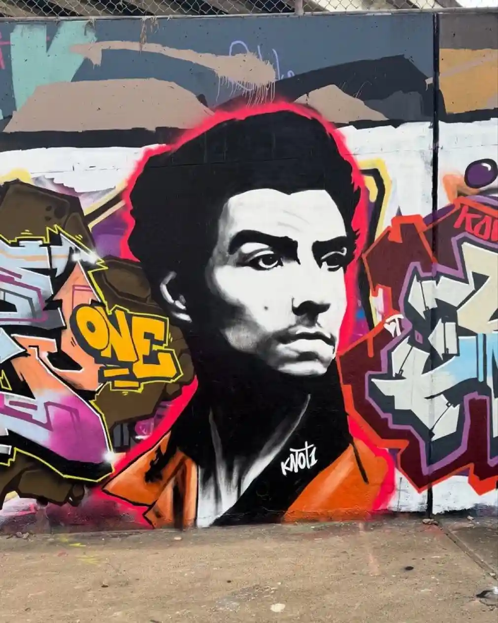 A black-and-white graffiti mural of a man's face with a red outline, set against a colorful, chaotic background of other…