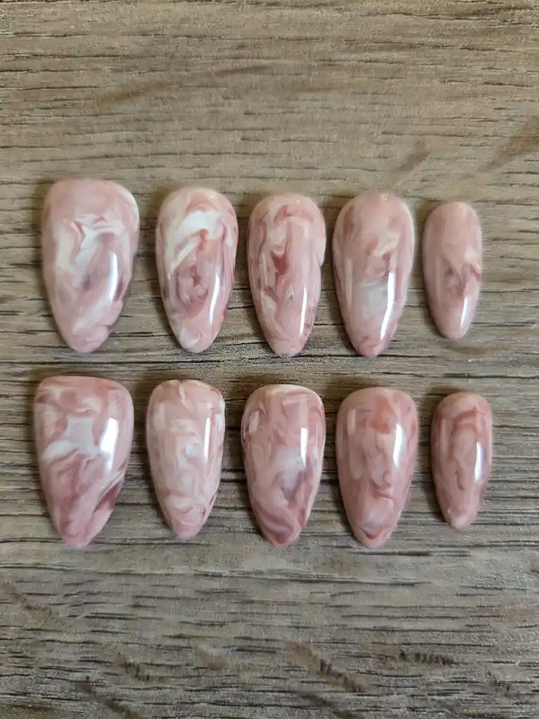 Ten artificial nails with a glossy pink and white marble pattern are arranged in two rows on a wooden surface. The nails are…
