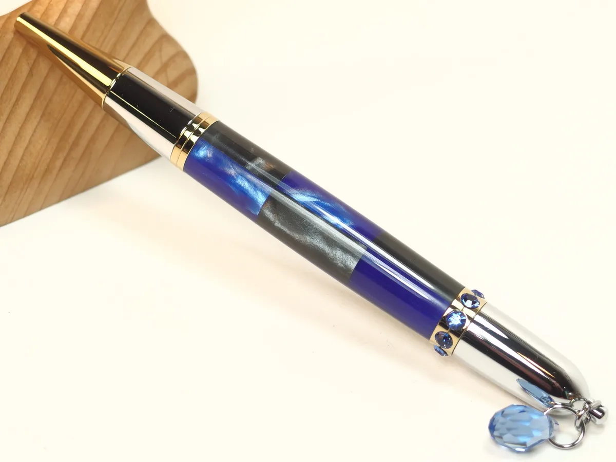 A highly decorated pen with gold, silver, and blue accents, adorned with blue crystals and a dangling crystal charm, rests…