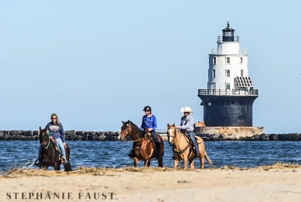 Three people riding horses on a beach with a lighthouse in the background. The rider on the right wears a cowboy hat and is…