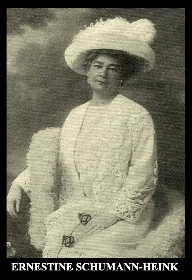 A vintage black-and-white portrait of a woman seated, wearing an ornate white lace dress and a large, fluffy white hat. The…