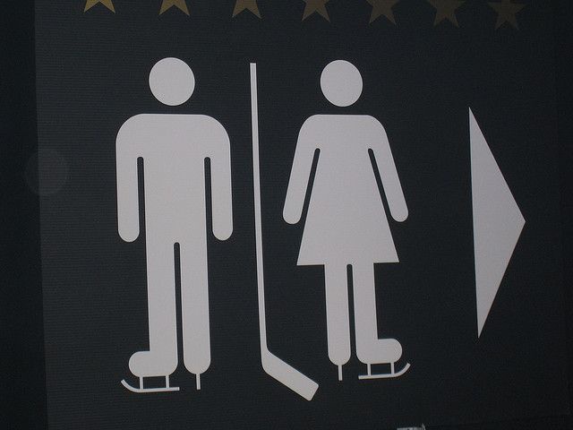 A dark sign with white pictograms shows a male ice hockey player holding a stick next to a female ice skater, both on…