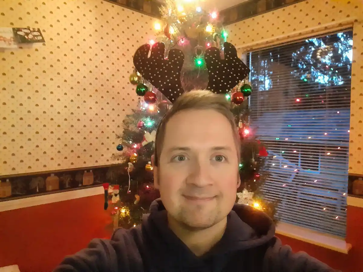 A man in a dark hoodie smiles at the camera while wearing a black polka-dotted reindeer antler headband. Behind him is a…