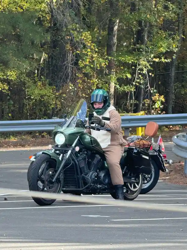 A person wearing a green helmet and tan outfit sits on a green motorcycle parked in a lot, looking down at a phone held in…