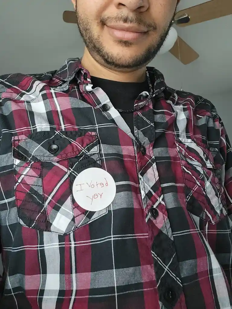 A person wearing a red, black, and white plaid shirt has a white circular sticker on their chest pocket. The sticker has red…