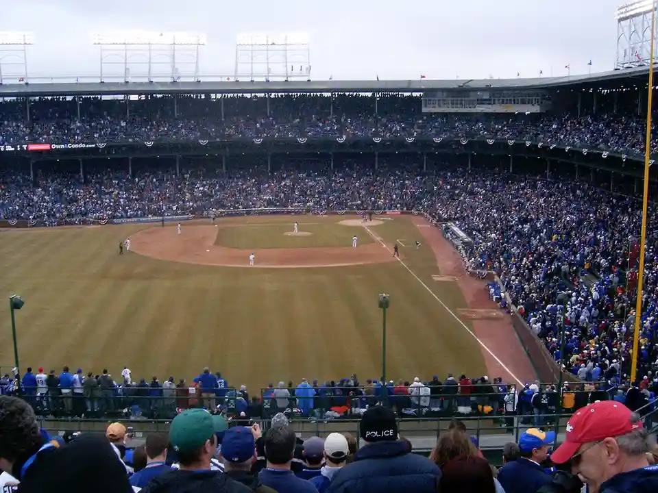 A wide shot of a crowded baseball stadium during a game, with players on the field and fans in the stands. In the…
